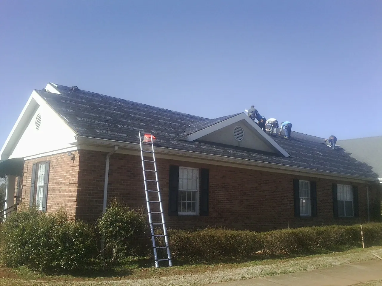 Crew tearing shingles off a brick ranch roof with a ladder set — Emergency Roof Repair work by our crew in Elmont