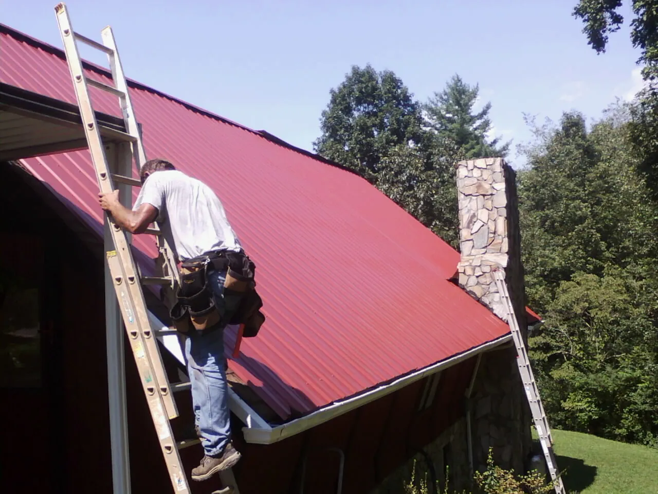Roofer on a ladder beside a red metal roof and stone chimney — Soffit & Fascia Repair by Roofing Brothers in Elmont